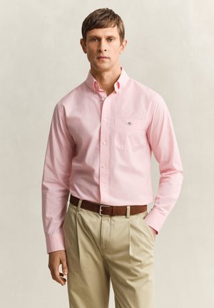 Homme portant une chemise à carreaux rose clair boutonnée et rentrée dans un pantalon beige avec une ceinture marron, debout devant un fond neutre.