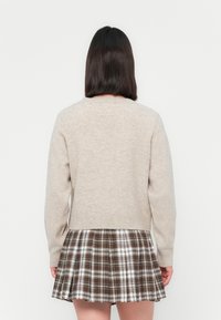 Beige kötött pulóver hosszú ujjakkal és bordázott aljjal, amely egy barna, fehér és szürke színű, bővülő, kockás szoknyával van párosítva.