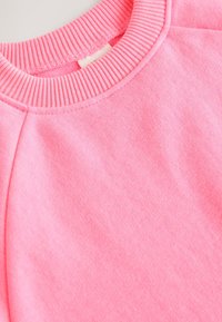 Zbliżenie na różową bluzę z prążkowanym okrągłym dekoltem, szwem na ramieniu oraz miękką, bawełnianą tkaniną.