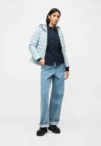 Γυναίκα που φοράει ανοιχτό μπλε μπουφάν puffer πάνω από μαύρο πουκάμισο με κουμπιά, φαρδιά ανοιχτό μπλε τζιν με γυρισμένα μπατζάκια στον αστράγαλο και μαύρα χοντρά παπούτσια, όρθια.