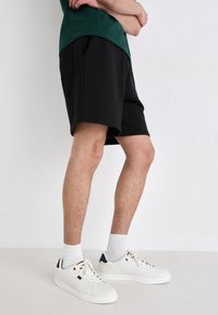 Pantalones cortos de algodón negros con un corte relajado, que cuentan con bolsillos laterales y una longitud ligeramente por encima de la rodilla, combinados con zapatillas blancas y calcetines.