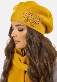 Béret en laine jaune avec deux accents floraux en beige et une étiquette de marque. Superposé avec une écharpe jaune assortie, présentant une texture accentuée.