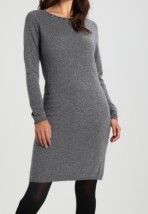Kobieta w dopasowanej szarej dzianinowej sukience z długim rękawem, z okrągłym dekoltem i subtelnymi zdobieniami, w połączeniu z czarnymi rajstopami.