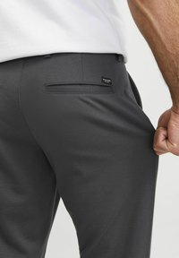 Pantalones grises con una textura suave, que presentan un bolsillo trasero y una pequeña etiqueta negra. La cintura es elástica y una mano sostiene la tela.