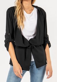 Chaqueta negra de estilo envolvente, hecha de tela suave, con mangas enrolladas y un lazo en la cintura. Se lleva sobre una camisa blanca y jeans de mezclilla.