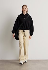 Felpa nera con coulisse in vita, abbinata a pantaloni beige a gamba larga. Porta una borsa a tracolla nera. Pavimento e sfondo sono neutri.