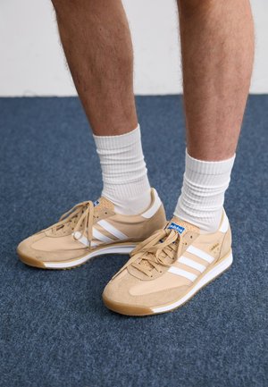 Béžové semišové tenisky s bielymi pruhmi a šnúrkami. Obsahujú textúrovaný zvršok, mäkkú podrážku a modrú štítok s logom na jazyku.