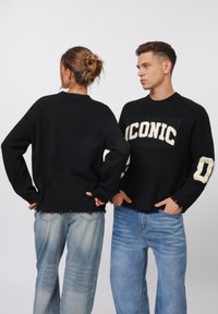 Black knit sweaters featuring “ICONIC” lettering in cream, with distressed hems. Paired with light blue, loose-fit jeans, showcasing denim texture.