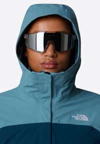 Blå og teal vandtæt jakke med høj krave og hætte, der har North Face logoet. Reflekterende oversized solbriller.
