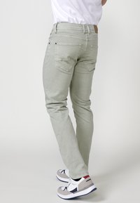 Jeans de mezclilla verde claro con corte slim, bolsillos traseros y parche de cuero en la cintura. Combinados con zapatillas grises y blancas con acentos rojos.