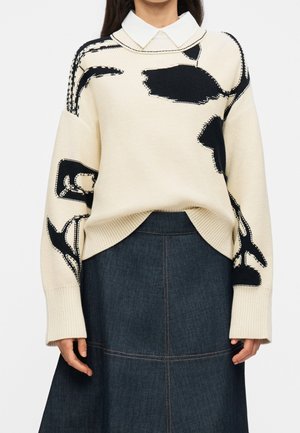 Femme portant un pull crème avec des motifs abstraits noirs sur une chemise blanche à col et une jupe en denim foncé.