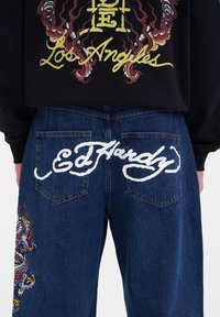 Denimjeans med blå tvätt, med broderad drake på vänster ben och vit text "Ed Hardy" på bakfickorna.