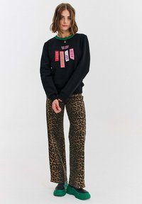 Černá mikina s barevnými textovými nášivkami na přední straně, spárována s hnědými kalhotami s leopardím potiskem a zelenými platformovými botami.