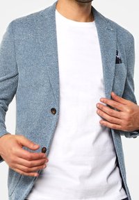 Ljusblå strukturerad kavaj med två bruna knappar, slag och en fickkvadrat, lager på en vit t-shirt. Skräddarsydd passform, mjukt tyg.