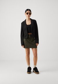 Svart cropped blazer, svart blonde topp, olivenfarget miniskjørt med glidelåsdetaljer, kombinert med svarte sneakers og brune sokker. Brune solbriller.
