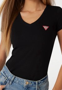 Femme portant un T-shirt noir à col en V et manches courtes avec un petit logo Guess rouge et blanc sur la poitrine, rentré dans un jean en denim bleu.