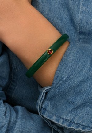 Bracelet vert brillant avec une surface lisse, présentant un accent doré avec une petite pierre précieuse rouge. Porté sur un poignet partiellement recouvert de denim.