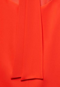 Rode blouse met een gladde textuur, voorzien van een brede, vloeiende strik bij de halslijn. De stof lijkt lichtgewicht en vloeibaar.