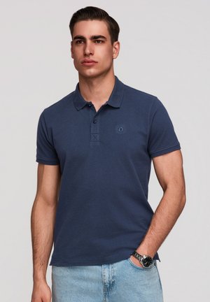 Jeune homme aux cheveux courts et foncés portant un polo bleu marine et un jean bleu clair, debout avec une main dans la poche.