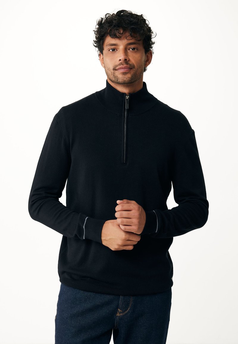 Pullover nero con mezza zip realizzato in tessuto morbido, caratterizzato da maniche lunghe, polsini a costine e una texture liscia. Indossato sopra pantaloni in denim.