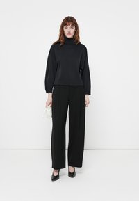 Schwarzer Rollkragenpullover mit weiten Ärmeln kombiniert mit schwarzen, hochgeschnittenen Hosen. Model hält eine kleine weiße Handtasche und trägt schwarze Schuhe.