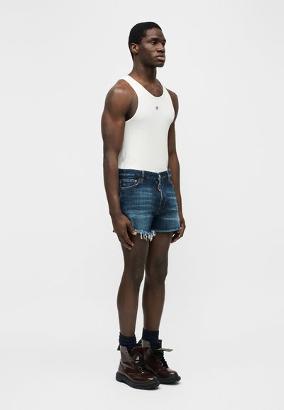 Μυώδης άτομο που στέκεται, φοράει λευκή αμάνικη μπλούζα, ξεφτισμένα μπλε τζιν σορτς, σκούρες κάλτσες και στιλβωμένα καφέ μποτάκια σε λευκό φόντο.