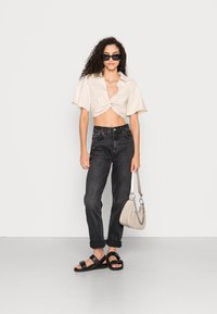 Camisa bege cortada com botões e um nó à frente, jeans pretos de cintura alta, sandálias pretas e uma bolsa cinza clara com alça de corrente.