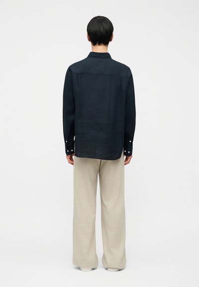 Άνδρας που φοράει μακρυμάνικο μαύρο πουκάμισο και φαρδιά μπεζ παντελόνια, στέκεται με τα χέρια χαλαρωμένα, γυρισμένος προς τα πίσω μπροστά σε ανοιχτόχρωμο απλό φόντο.