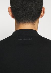 Bakvy av en person som bär en svart tröja med "KARL LAGERFELD" präglat på kragen på övre ryggen.