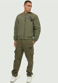 Olivno zeleni prešiti bomber jakna z zadrgo in velikim logotipom, v kombinaciji z usklajenimi cargo hlačami, ki imajo stranske žepe in blagovno znamko.