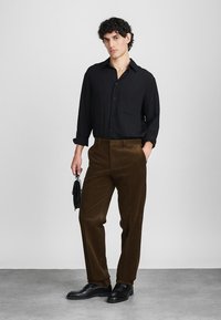 Camicia nera con bottone e vestibilità rilassata, abbinata a pantaloni in corduroy marroni. Il modello tiene una piccola borsa nera e indossa scarpe nere.