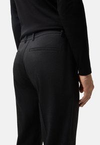 Pantalon noir ajusté avec une texture lisse, des passants de ceinture et une poche arrière. Design ajusté, présenté de dos avec une chemise ajustée.