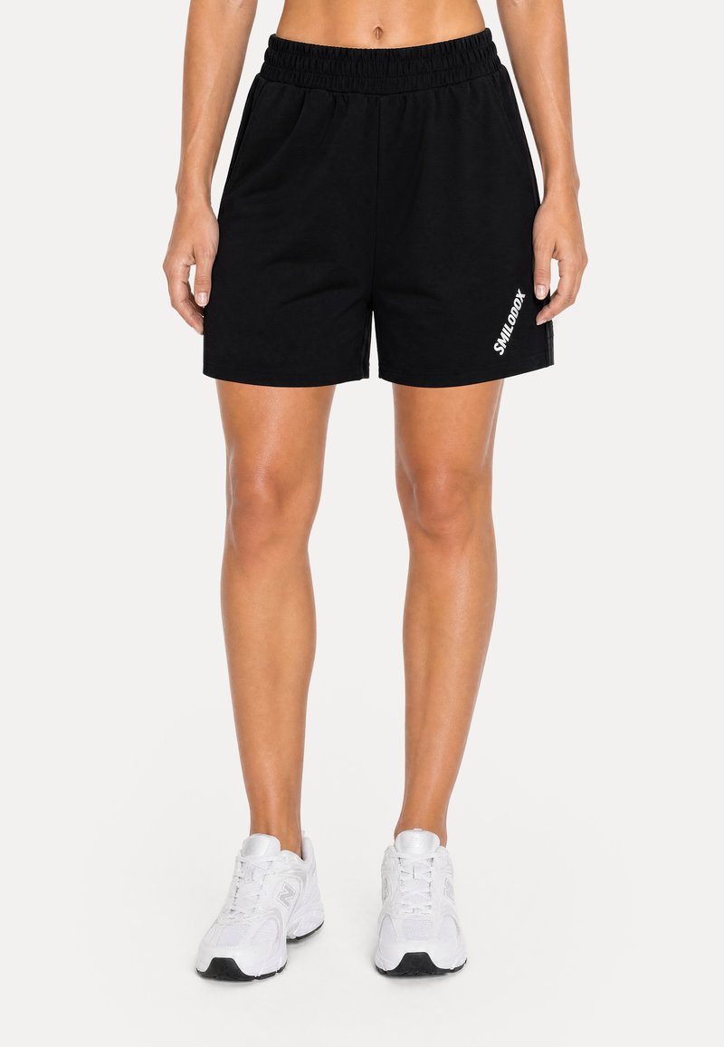 Personne portant un short noir à taille élastique avec un texte blanc "SMILODOX" sur la jambe gauche et des chaussures de sport blanches, debout sur un fond blanc.