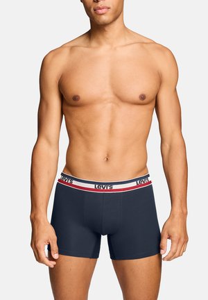 Oben ohne Mann, der marineblaue Levi's Boxershorts mit einem weißen, roten und marineblauen Bund trägt, steht vor einem schlichten hellen Hintergrund.
