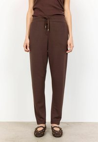 Bruine joggingsbroek van zacht materiaal, met een tailleband met trekkoord, taps toelopende pijpen en zijzakken. Textuurrijke bruine ballerina's maken de look compleet.