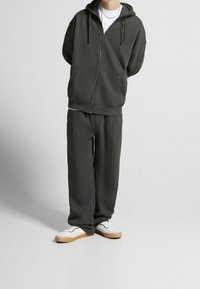 Sudadera con cremallera de color verde oscuro, con bolsillos tipo canguro y pantalones a juego. La tela parece suave y tiene un corte relajado; se lleva con zapatillas blancas.