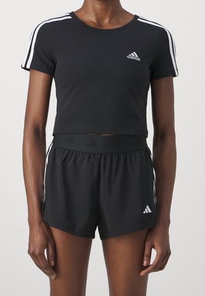 Osoba nosząca czarną przyciętą koszulkę Adidas i czarne sportowe szorty Adidas z białymi paskami na rękawach i bokach szortów.