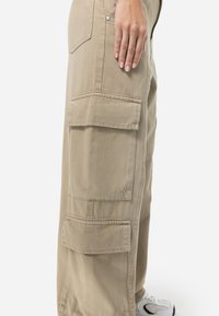 Beige cargo bukser laget av bomull, med store sidelommer, en avslappet passform, og en synlig metallknapp på livet.