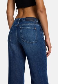Blaue Jeans mit hoher Taille und geradem Bein, versehen mit Gesäßtaschen und einem Lederetikett am Bund. Texturiertes Gewebe mit leichtem Glanz.