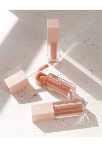 Drie doorzichtige lipgloss-buizen met lichtroze doppen. Twee bevatten getinte gloss, één heeft een applicator. Gladde texturen en minimalistisch ontwerp.