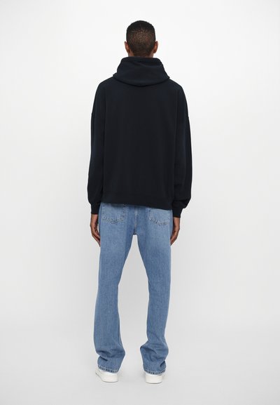 Schwarzer Kapuzenpullover und hellblaue, locker sitzende Jeans mit einfachem Design, elastischen Bündchen und Gesäßtaschen, vor einem neutralen Hintergrund.