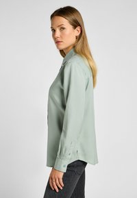 Chemise vert clair à col en tissu lisse, avec des manches longues, des poignets boutonnés et un ourlet arrondi. Portée avec un jean foncé.