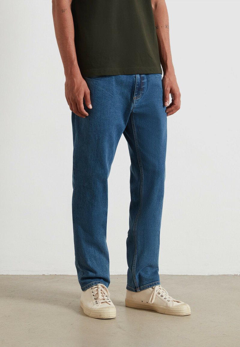 Jeans in denim blu con un design a gamba dritta, caratterizzati da una vita media e cuciture sottili. Indossati con sneakers in tela beige.