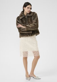 Barna műszőrme dzseki magas gallérral, krémszínű gyöngyökkel díszített csipkerózsa szoknyával és ezüst színű, hegyes orrú, nyitott sarkú cipővel párosítva.