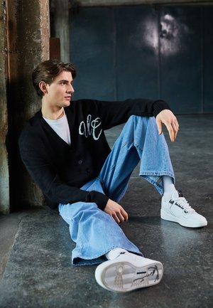 Cardigan noir avec broderie blanche "ale", t-shirt blanc, jean large bleu clair, et baskets blanches avec accents noirs. Fond industriel.