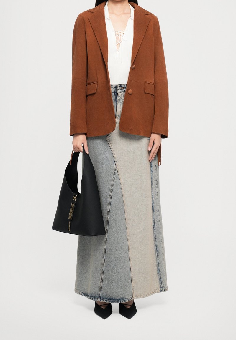Femme portant un blazer marron, un haut blanc à lacets, une longue jupe en jean bicolore, des chaussures noires pointues, tenant un sac à main noir.
