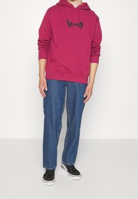 Felpa con cappuccio bordeaux con logo nero, tasca a marsupio frontale, abbinata a pantaloni in denim blu e sneakers nere. Vestibilità casual, materiale in cotone.
