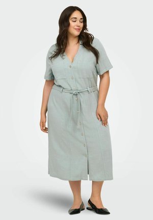 Kvinde med langt mørkt hår iført en lyseblå midi-kjole med knapper og bælte i taljen samt sorte slingback-sko, stående og kiggende ned.