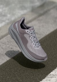 Lyse grå treningssko med et teksturert mesh-overdel, rund tå og fleksibel såle, med markante stripe mønstre og snørebånd.