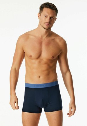 Oberkörperfreier Mann mit kurzen Haaren, der marineblaue Boxershorts mit einem blauen Bund trägt, auf dem "Schiesser" steht, vor einem schlichten weißen Hintergrund.